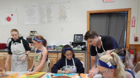 Counselor demonstrates cutting skills to a group of campers wearing aprons and seated around a table.