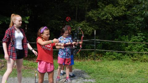 Youth stand on firing line with bows drawn as counselor looks over them.