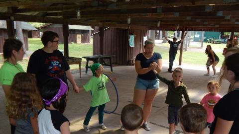 Day campers in a circle holding hands and passing a hula hoop around as a counselor times them with a stopwatch.