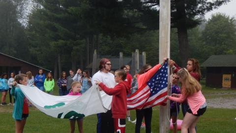 Campers work together with a counselor to raise the American and 4-H flags as the rest of camp looks on.
