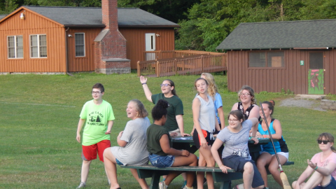 Cabin group seated at a picnic table with counselor who is waving arm.