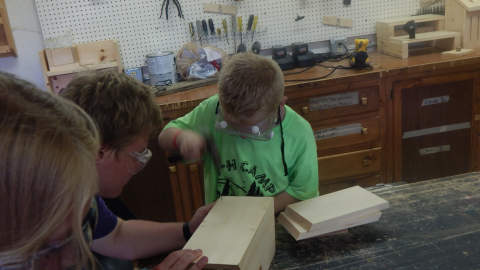 Counselor holds a wood project steady for a camper using a hammer.