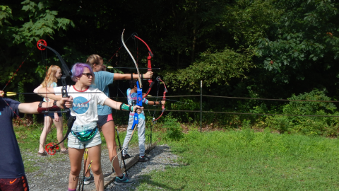 campers standing in a line with bows drawn as a counselor walks behind them.