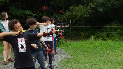 youth stand on the shooting line with bows drawn and a ground quiver of arrows in a ground quiver beside them while a counselor watches over them.