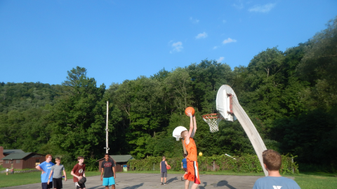 camper jumping up to dunk a basket on the bball court while others watch.