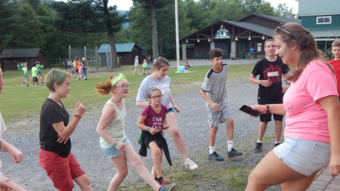 Counselor demonstrates a dance for campers who follow along kicking their feet and laughing.