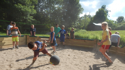 Youth hits a rubber ball in the gaga pit as others scatter behind them so as not to be hit. Players who have been eliminated watch from outside the gaga pit.