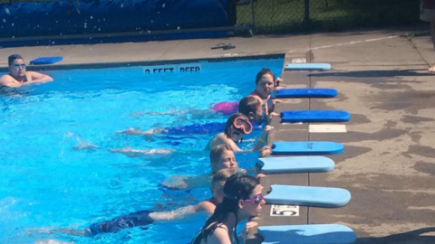 Youth hold the side of the pool with legs outstretched kicking. Each youth has a kickboard in front of them on the pool deck.