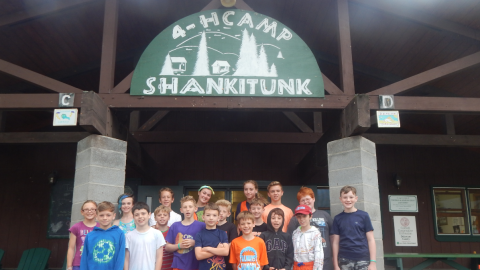 Group of 16 youth gathered together under the camp dining hall sign.