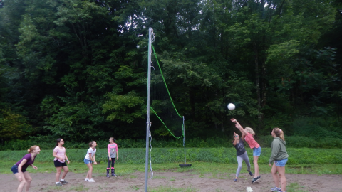 youth playing volleyball. One youth jumps toward the ball on the right side of the net with arms extended after hitting it toward the opposing team of four.